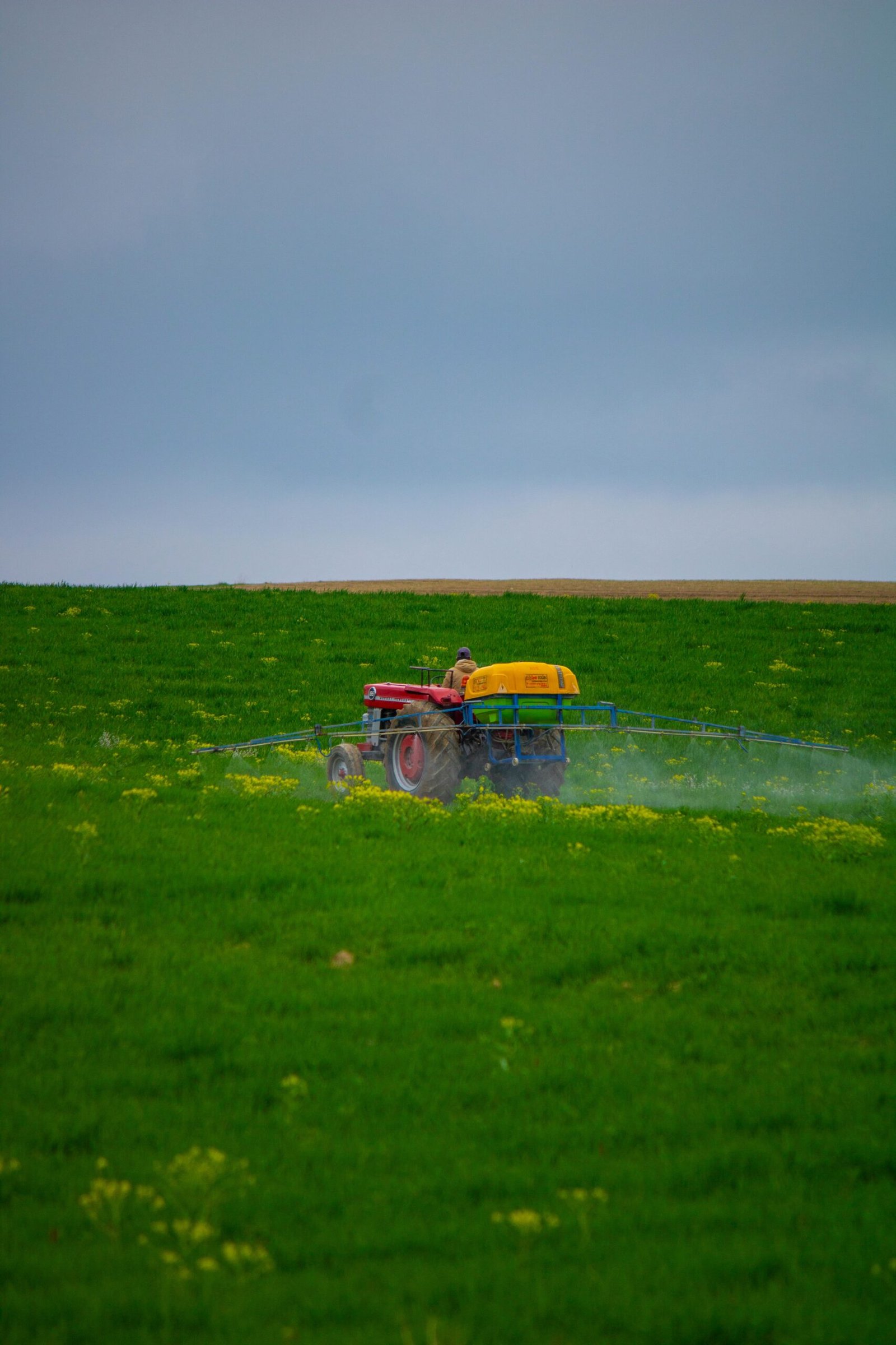 A farmer in a tractor spraying pesticides on a lush green field under a cloudy sky.